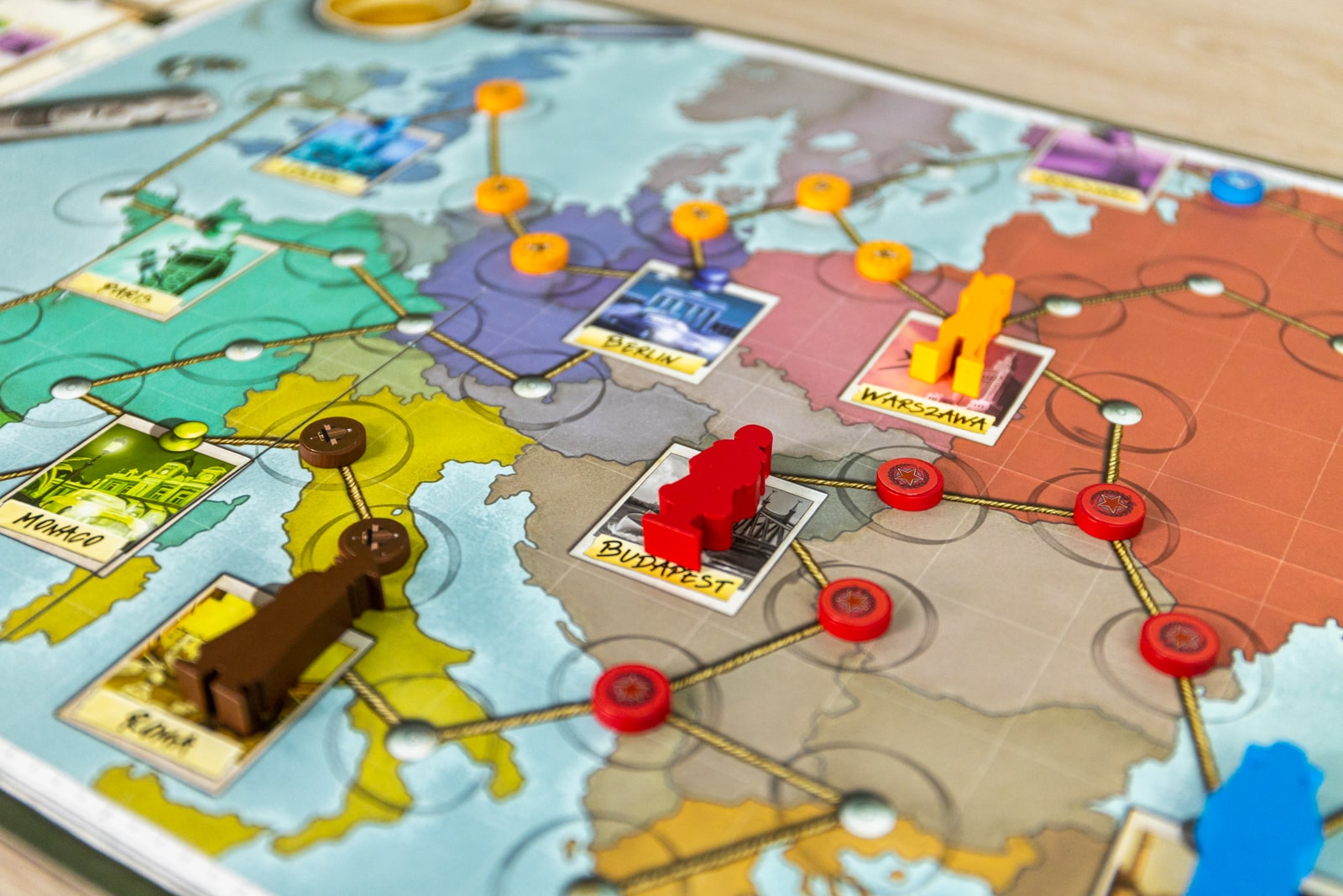 A close-up of the Spy Connection board game map of Europe by Pegasus Spiele shows city names and routes, with colored player tokens and red discs on cities like Warsaw, Berlin, Budapest, and Rome.