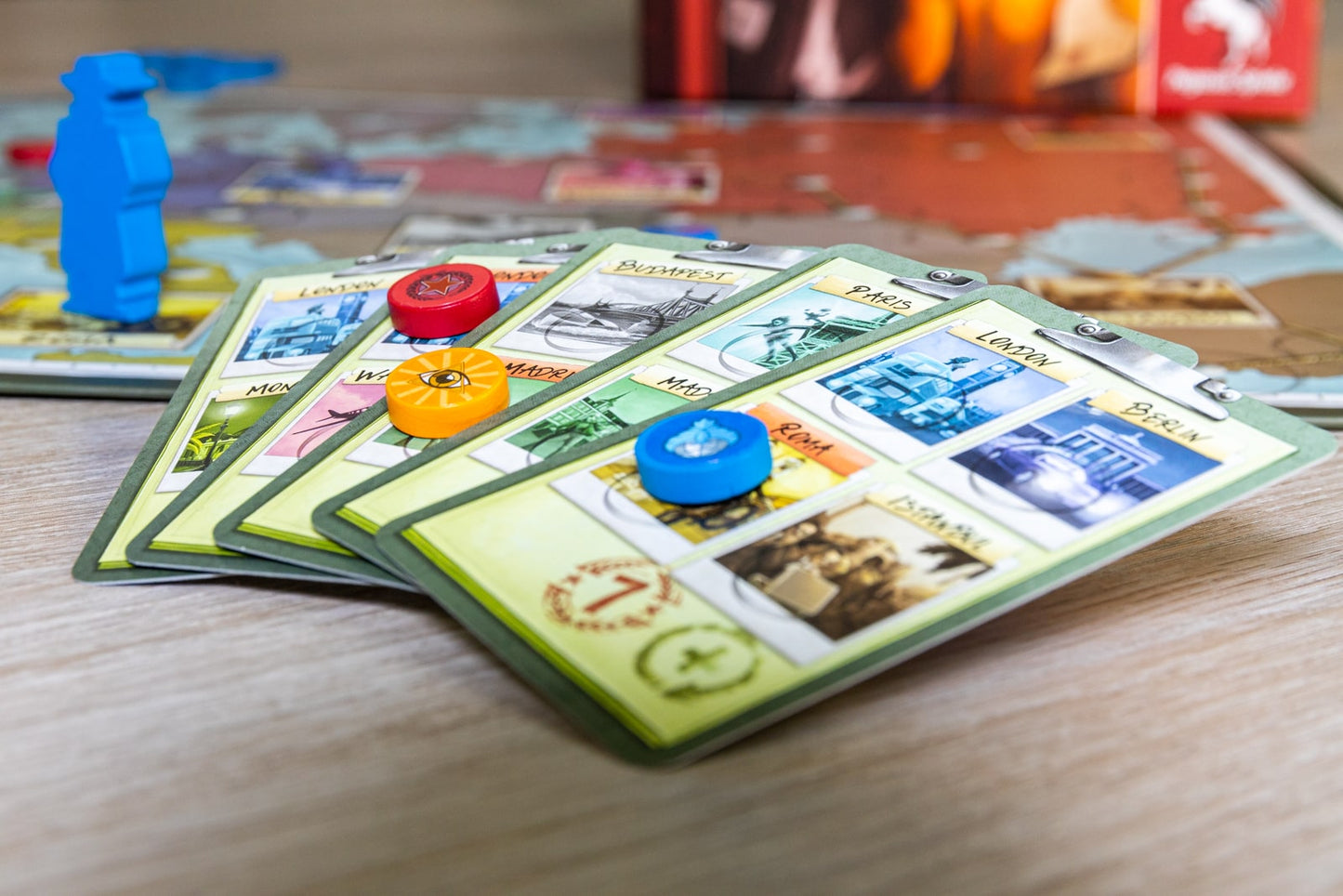 A close-up of five colorful Spy Connection cards from Pegasus Spiele, each showing different city names and tokens, with a blurred game board and blue pieces visible in the background.
