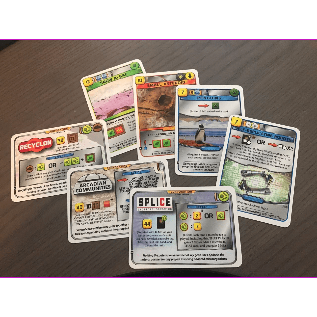 Seven illustrated cards from the Terraforming Mars: Promo Pack by Fryx Games are spread out on wood, each displaying unique science and technology-themed images, icons, numbers, and game instructions.