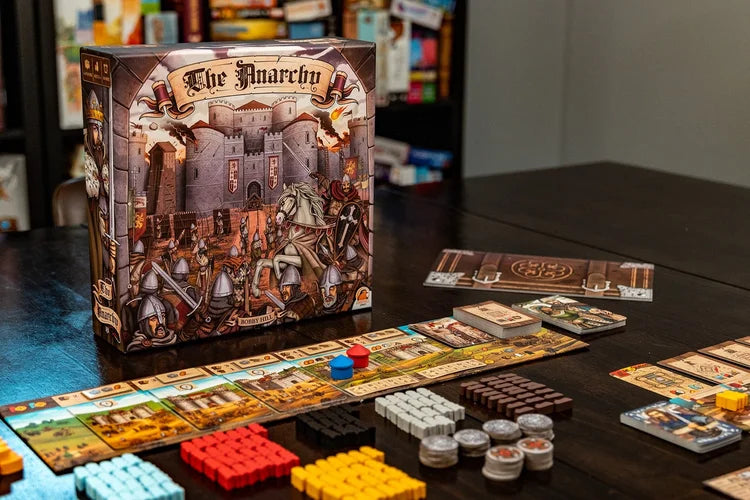 A tabletop setup for The Anarchy by Renegade Game Studios, showing the game box with medieval artwork and an array of colorful pieces, cards, and tiles neatly arranged on a dark table.