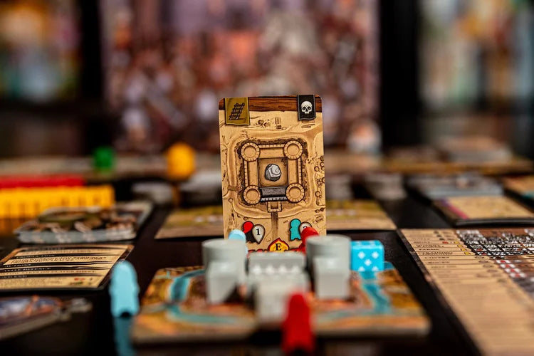 A close-up of The Anarchy by Renegade Game Studios shows its setup with cards, colorful pieces, gray castle tokens, and a blue die around a central castle card in focus. The background is blurred.