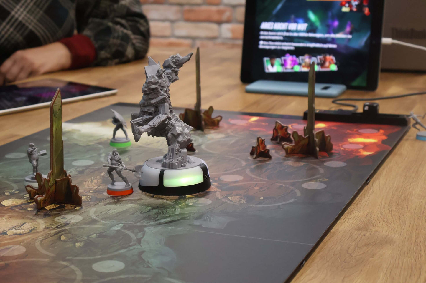 A player’s arm rests beside detailed miniatures on a 3D board for The Bad Karmas and The Curse of the Zodiac by Pegasus Spiele, while a digital screen shows game info in this cooperative tabletop experience.