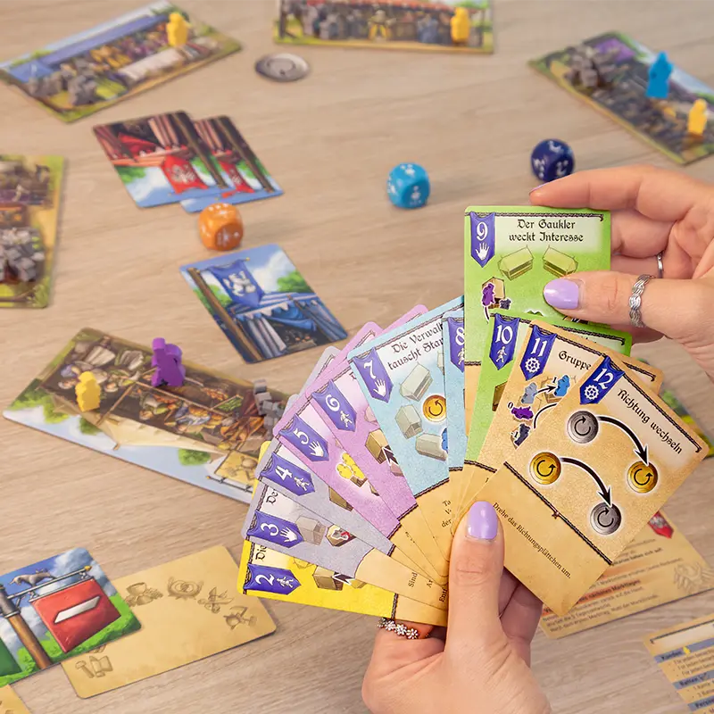 A player holds colorful influence cards while playing Tricky Traders by Pegasus Spiele, with game cards, dice, and pieces scattered on a wooden table in the lively setting of this marketplace board game.