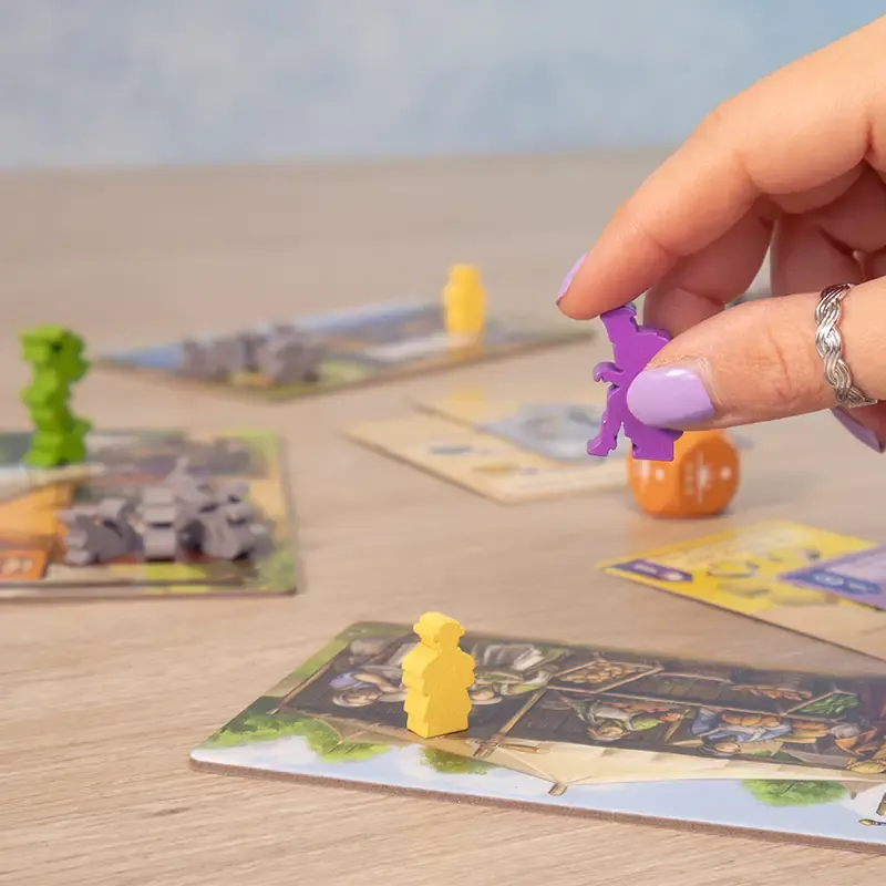 A hand with purple-painted nails moves a purple game piece on the Tricky Traders board game by Pegasus Spiele, surrounded by colorful pieces, influence cards, and a die on a wooden surface.