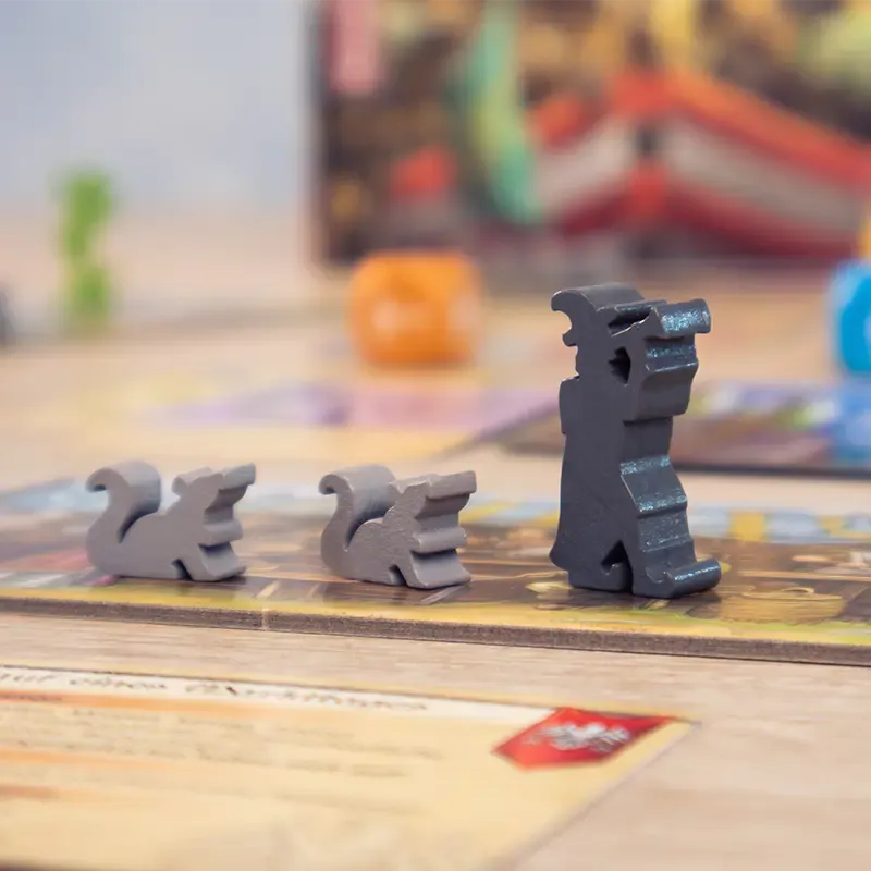 Three gray wooden dragon-shaped game pieces from Tricky Traders by Pegasus Spiele are on the board, with influence cards and other colorful components blurred in the background.