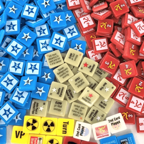 A close-up of GMT Games' Twilight Struggle Deluxe Plastic Pieces shows blue tokens with white stars, red ones with yellow hammer and sickle symbols, tan instructions, and yellow tiles with nuclear symbols and text.