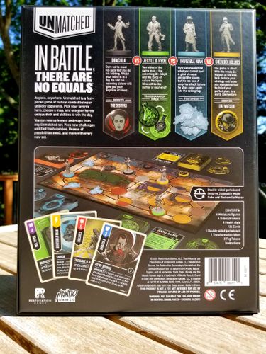 The back of the Unmatched: Cobble & Fog box from Restoration Games shows game components—character cards (Dracula, Jekyll & Hyde, Invisible Man, Sherlock Holmes), miniatures, and a description. The box rests on a wooden outdoor table.