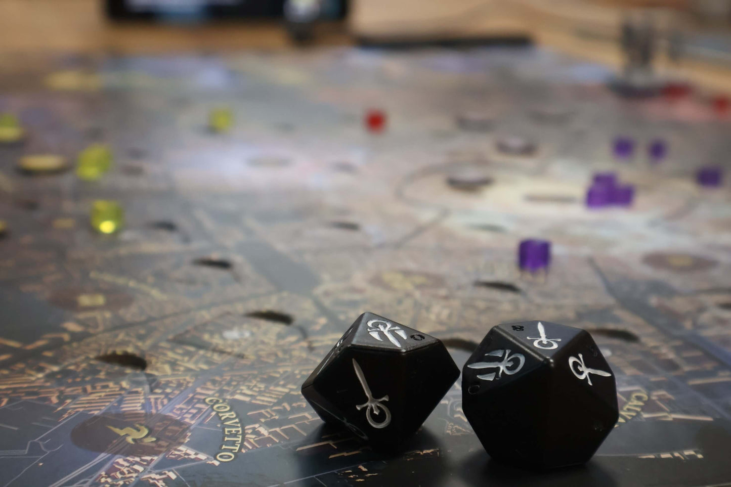 Two black twelve-sided dice with white markings rest on the detailed Teburu Board from Vampire: The Masquerade – Milan Uprising by Pegasus Spiele, surrounded by translucent colored game pieces and map-like designs, evoking a mysterious atmosphere.