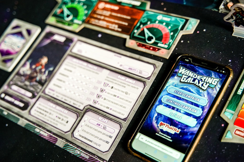 A smartphone displays the Wandering Galaxy: A Crossroads Game menu by Plaid Hat Games, lying beside game cards and character sheets on a space-themed table for an immersive sci-fi board game experience.