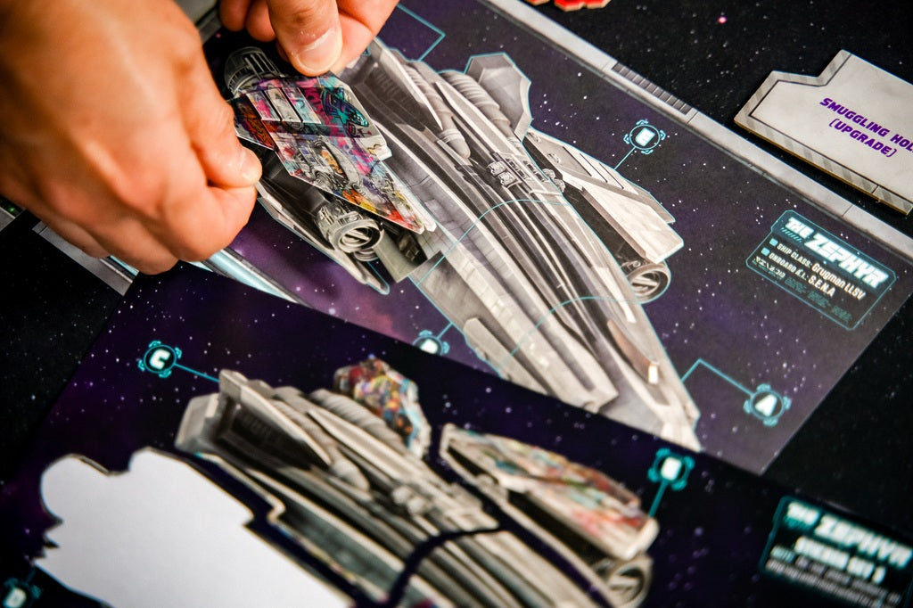 Close-up of hands assembling colorful cardboard spaceship pieces on Plaid Hat Games’ Wandering Galaxy: A Crossroads Game, with the sci-fi themed board, diagrams, and instructions visible.