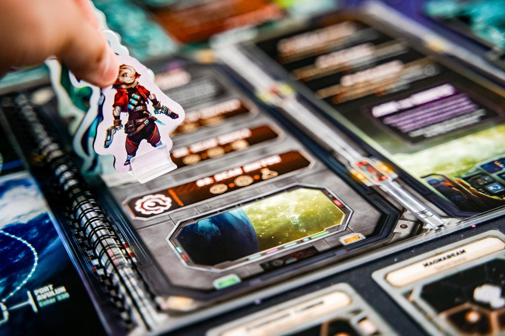 A hand holds a standee over the vibrant board of Plaid Hat Games’ "Wandering Galaxy: A Crossroads Game," featuring sci-fi art, cards, and text that suggest an epic space exploration adventure.