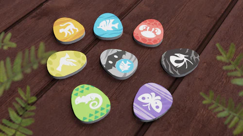 Eight colorful, egg-shaped tokens with animal silhouettes from Bad Comet's Wondrous Creatures sit on a wooden surface among green fern leaves—ideal for strategic board games with worker placement and tableau-building mechanics.