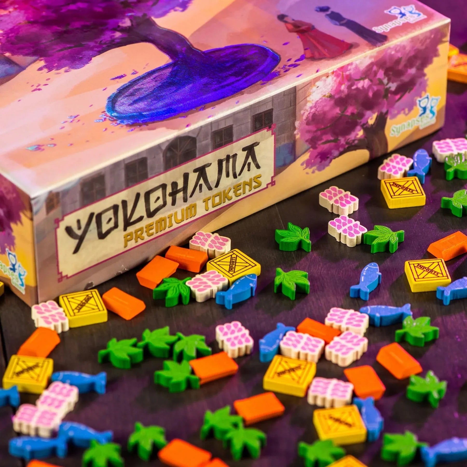 A box of Synapses Games' Yokohama: Premium Tokens sits on a table, surrounded by vibrant barrel, crate, fish, leaf, and tile tokens in orange, green, blue, yellow, and pink.