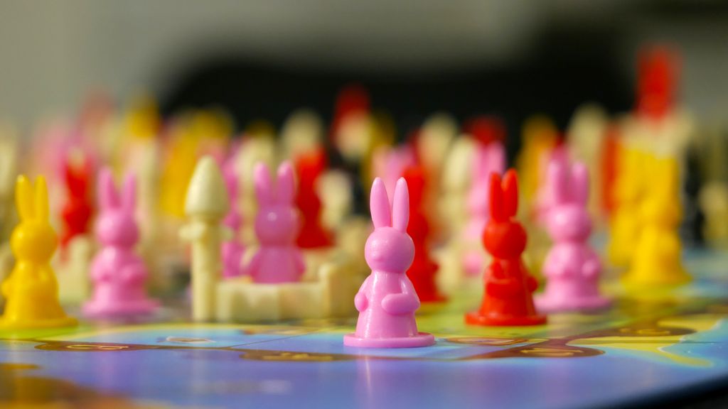 Pink, red, and yellow plastic rabbit clan pieces from Iello's Bunny Kingdom sit on a round board, with a pink rabbit in sharp focus at the front and other colorful rabbits blurred in the background.