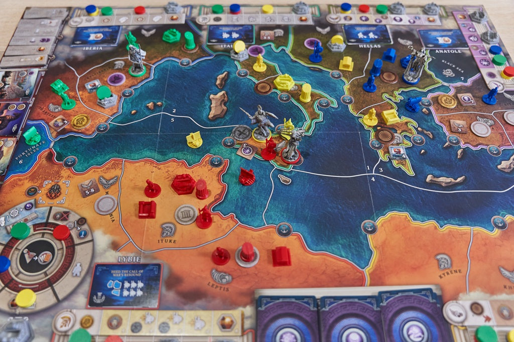 Pillars of Heracles by Apeiron Games is set up, showcasing tokens, miniatures, and cards on a detailed Archaic Period Greece map. Red, yellow, blue, and green city-state pieces are spread across the board.