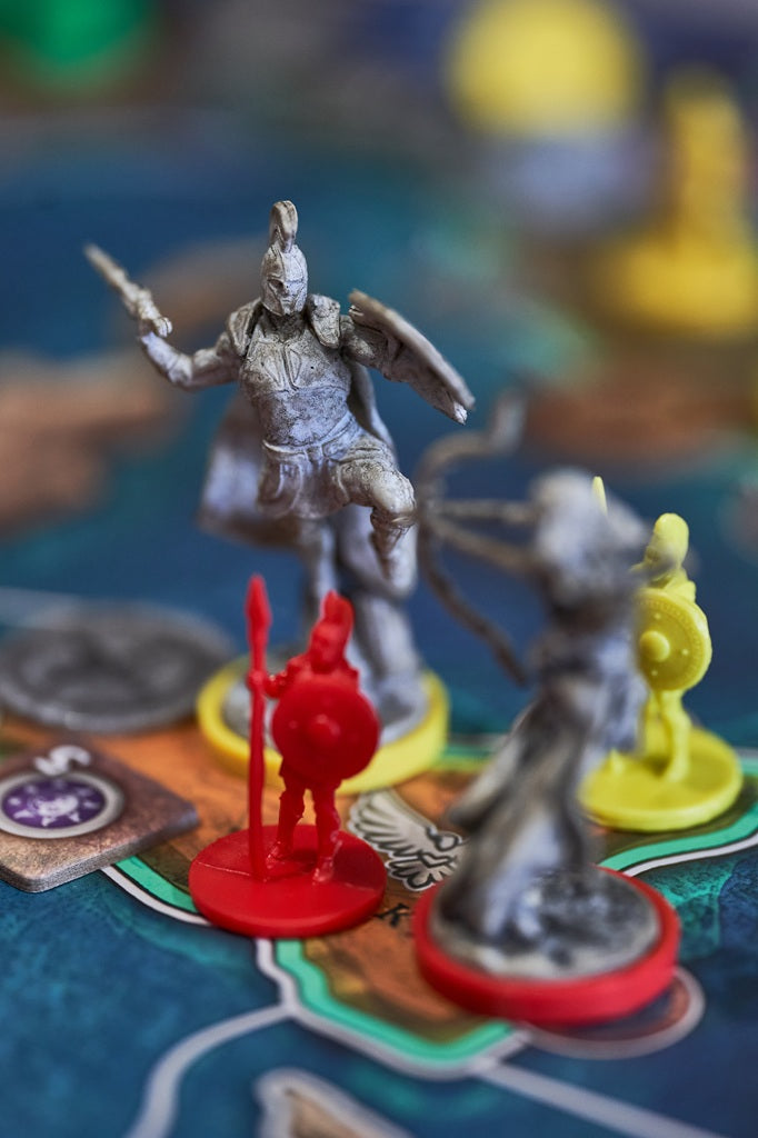 Close-up of four colorful miniatures from the "Pillars of Heracles" board game by Apeiron Games, featuring a red soldier and three silver and yellow warriors on a vibrant, ancient Greece city-state themed game board.