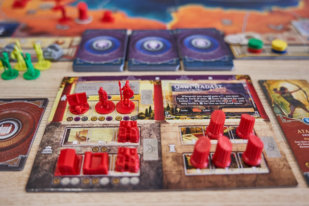 A close-up of Pillars of Heracles by Apeiron Games in play, with red figurines and tokens on illustrated city-state cards, green and yellow pieces in the background, and three circular design cards at the top center.
