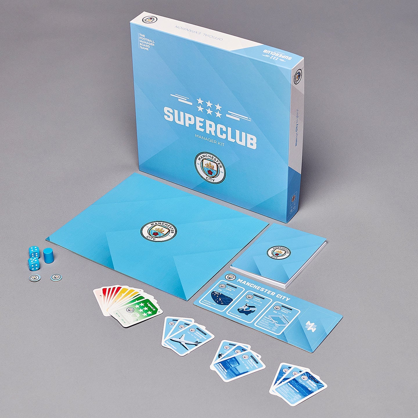 The Superclub: Manchester City Manager Kit by Superclub includes a game box, blue envelope, scorepad, blue dice, cards, player markers, and game boards—perfect for aspiring Man City managers. All items are arranged neatly on a gray surface.